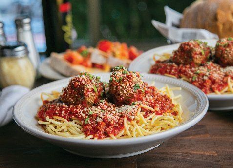 Spaghetti with meatballs, covered in red sauce and parmesan cheese, on white plates. A bread basket and bruschetta are in the background.