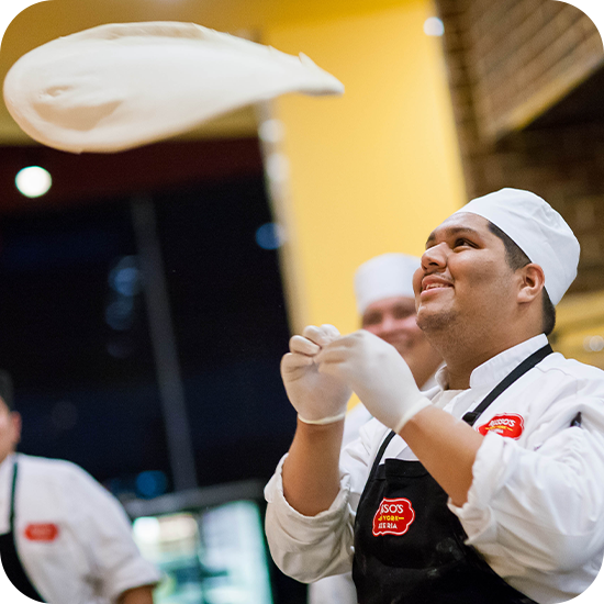 Chef tossing pizza dough in the air, smiling, wearing a chef hat, gloves, and apron.
