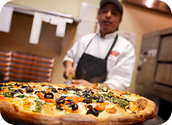 Pizza maker slicing a large pizza with olives, tomatoes, and greens.