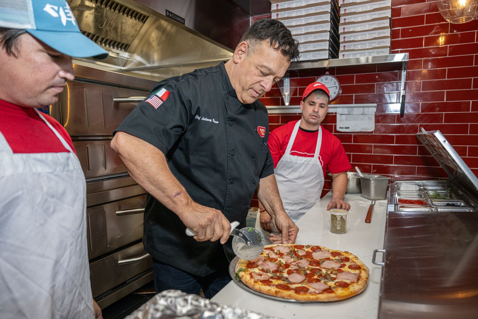 Chef cutting pizza in a kitchen, with two other people watching. Red brick wall.