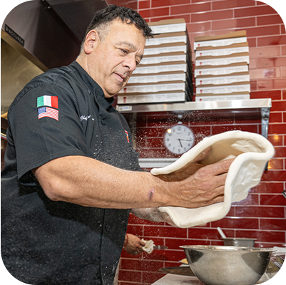 Chef tossing pizza dough in a restaurant kitchen with red brick walls.
