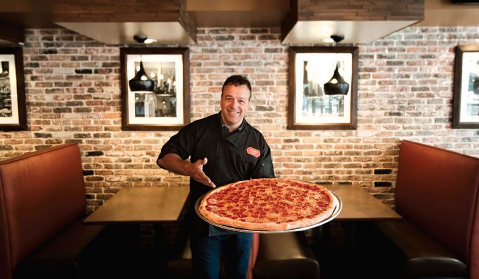 Chef in black uniform smiling, holding a large pepperoni pizza in a restaurant booth setting with brick walls and framed artwork.