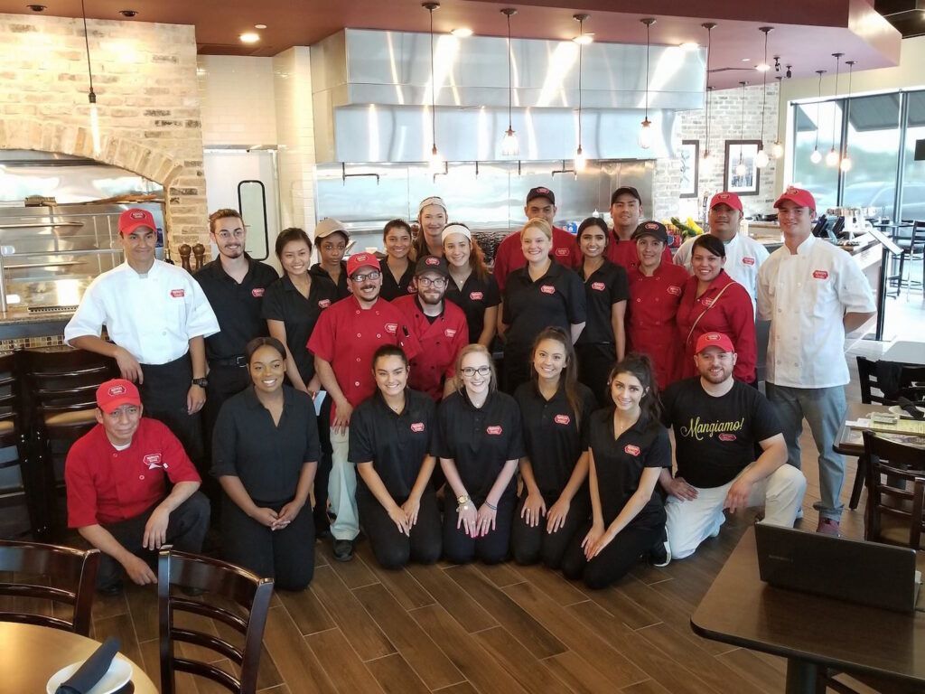 A group of restaurant employees poses for a photo in their restaurant. Many are wearing red shirts and hats, some black.