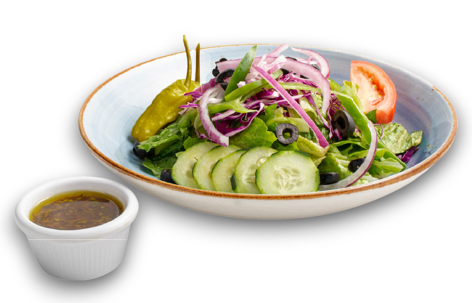 A colorful house salad with cucumbers, red onions, and a pepperoncini in a shallow bowl with a side of dressing.