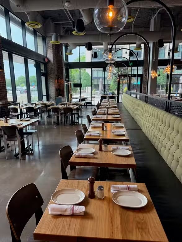 Modern restaurant interior with long wooden tables, cushioned bench seating, and large windows.