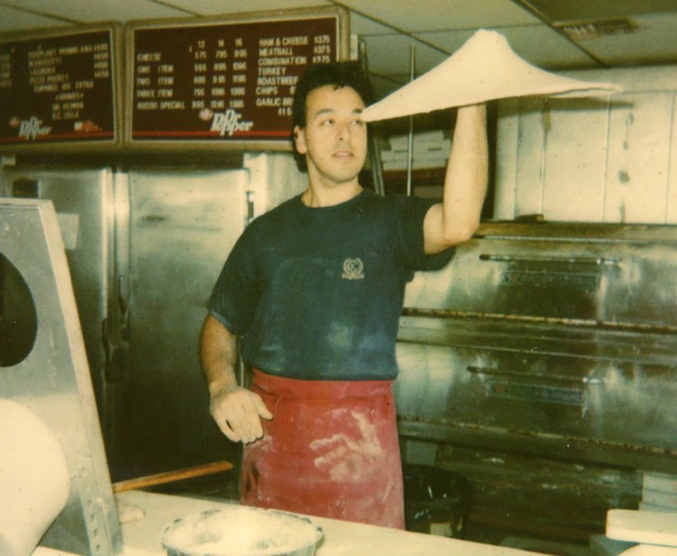 A man tossing pizza dough high in the air. He wears a red apron and looks towards the dough in a pizzeria.