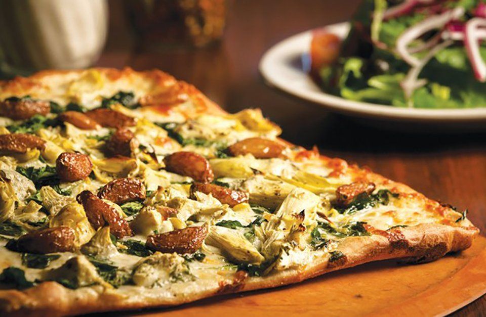Pizza topped with artichoke hearts, spinach, and sausage on a wooden board; a salad in the background.