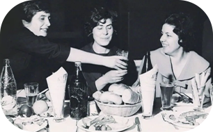 Three women at a table. One raises a glass, others smile. Bottles, food, and glasses visible.