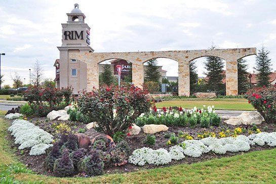 Entrance to RIM shopping center with a stone archway, flowerbeds, and a cloudy sky.