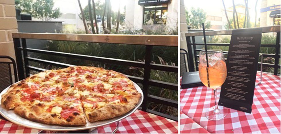 A pizza on a checkered table with a drink and menu on a patio. The setting has a railing with a view of trees.