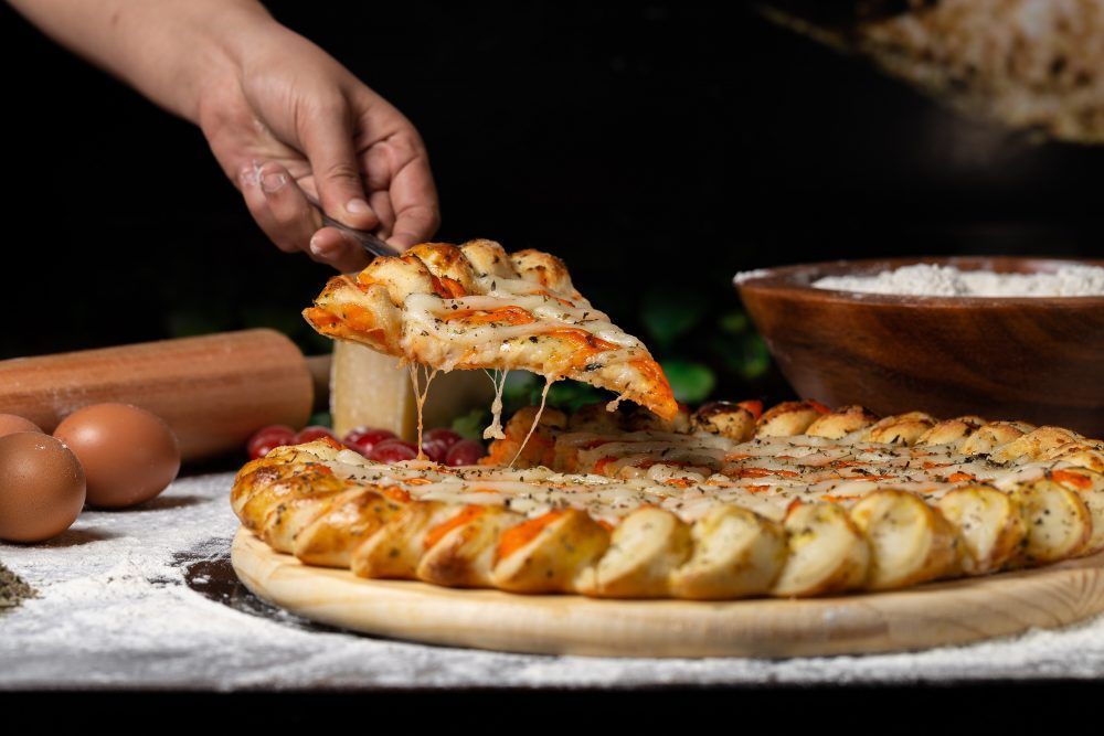 Hand lifting a slice of cheesy, braided-crust pizza from a wooden board, with ingredients like eggs and flour in the background.