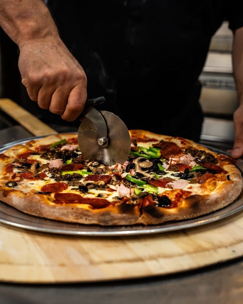 Person cuts a pizza with various toppings using a pizza cutter on a metal tray.