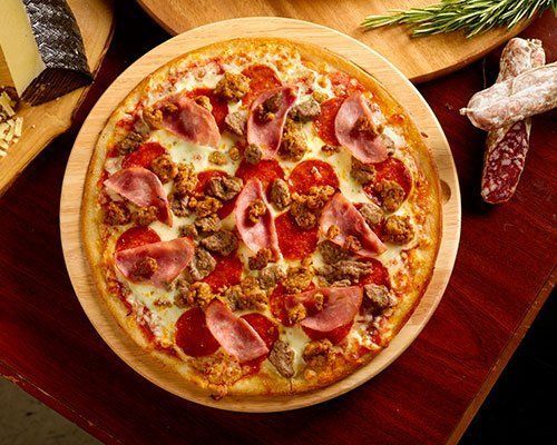 Pizza with pepperoni, ham, and sausage on a wooden pizza peel, alongside cheese and salami.