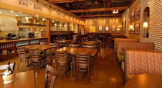 Restaurant interior with wooden tables, chairs, and booths; a brick wall, and an open kitchen with a 