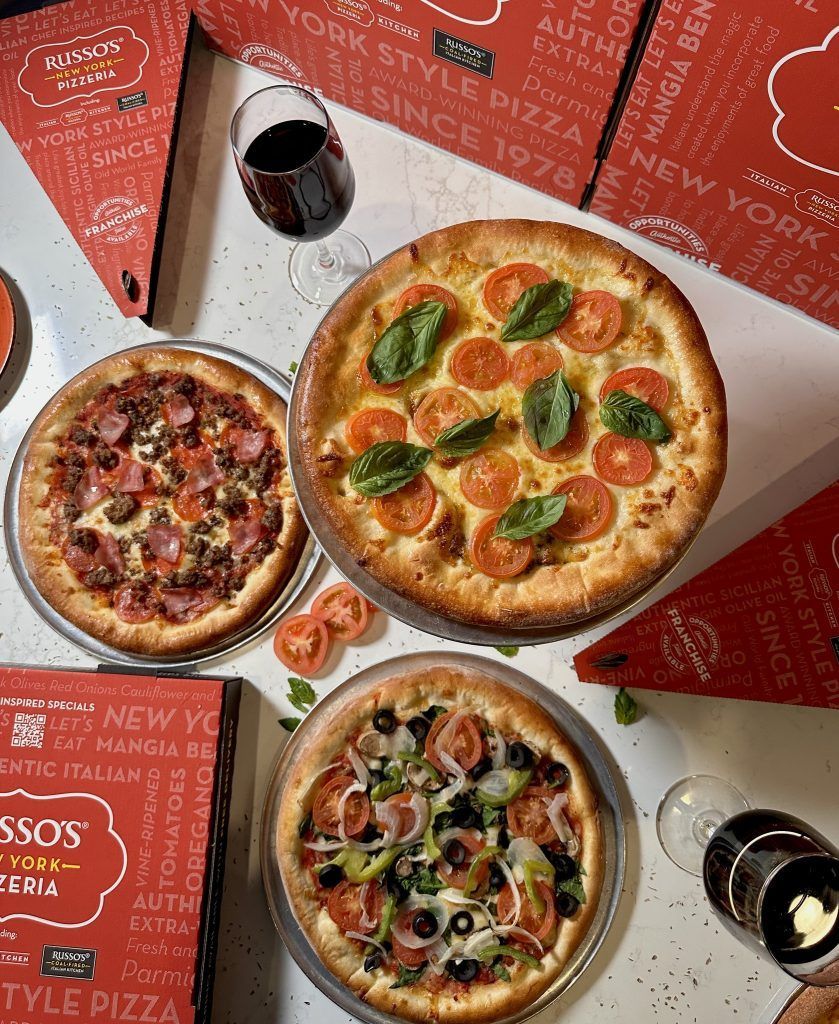 Three pizzas with various toppings, near pizza boxes, wine glasses and a bottle of wine.