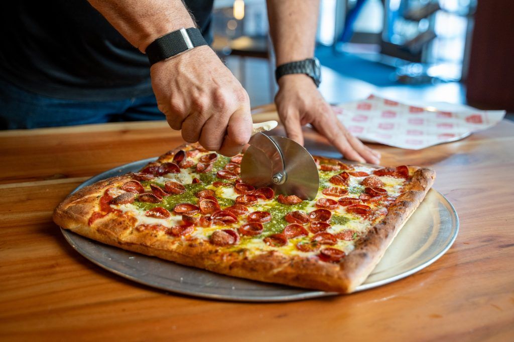 Person slicing a square pizza with a pizza cutter on a metal tray, wooden table.