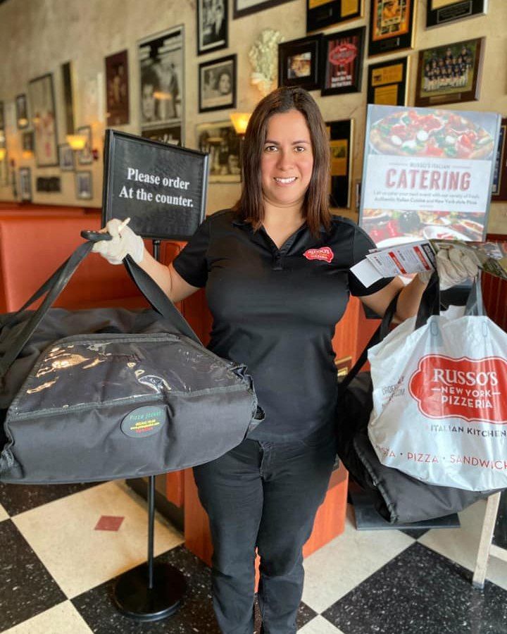 Woman holding delivery bags at Russo's, wearing a black shirt and pants, standing inside restaurant.