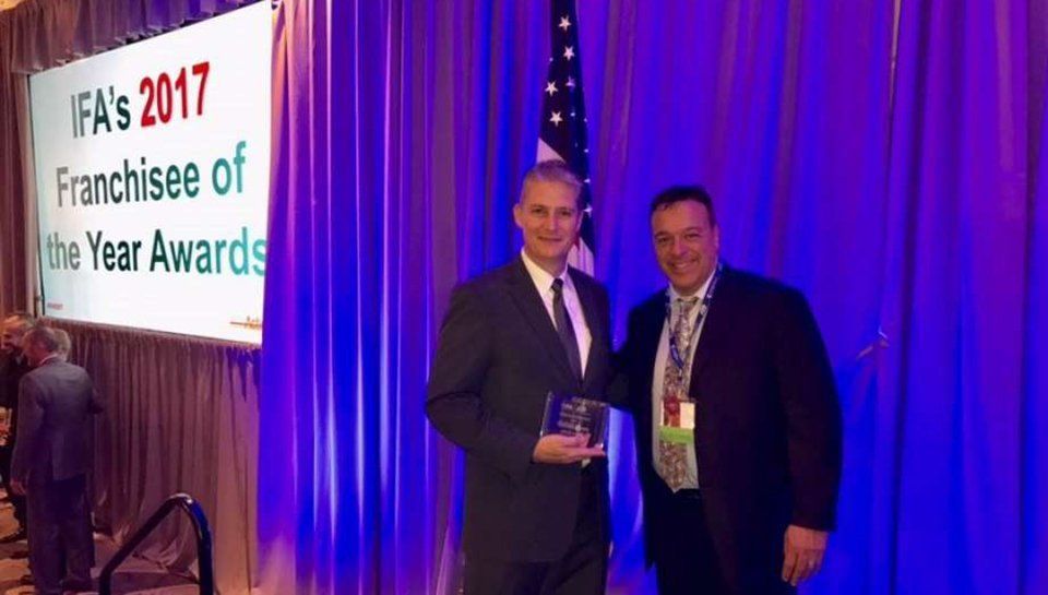 Two men in suits at an awards ceremony: one holding a trophy, both smiling in front of a screen that reads 