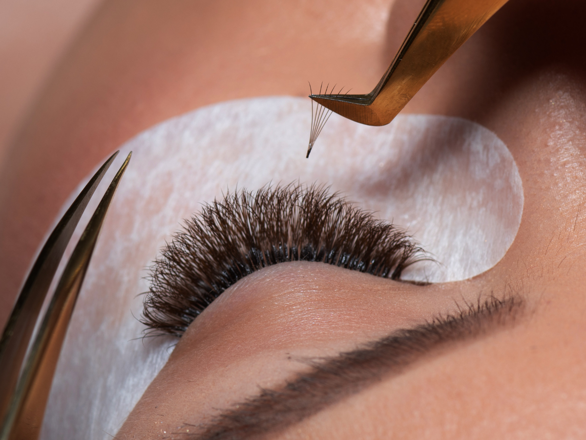 Eyelash extensions being applied; tweezers holding a lash cluster near a closed eye with a protective pad.