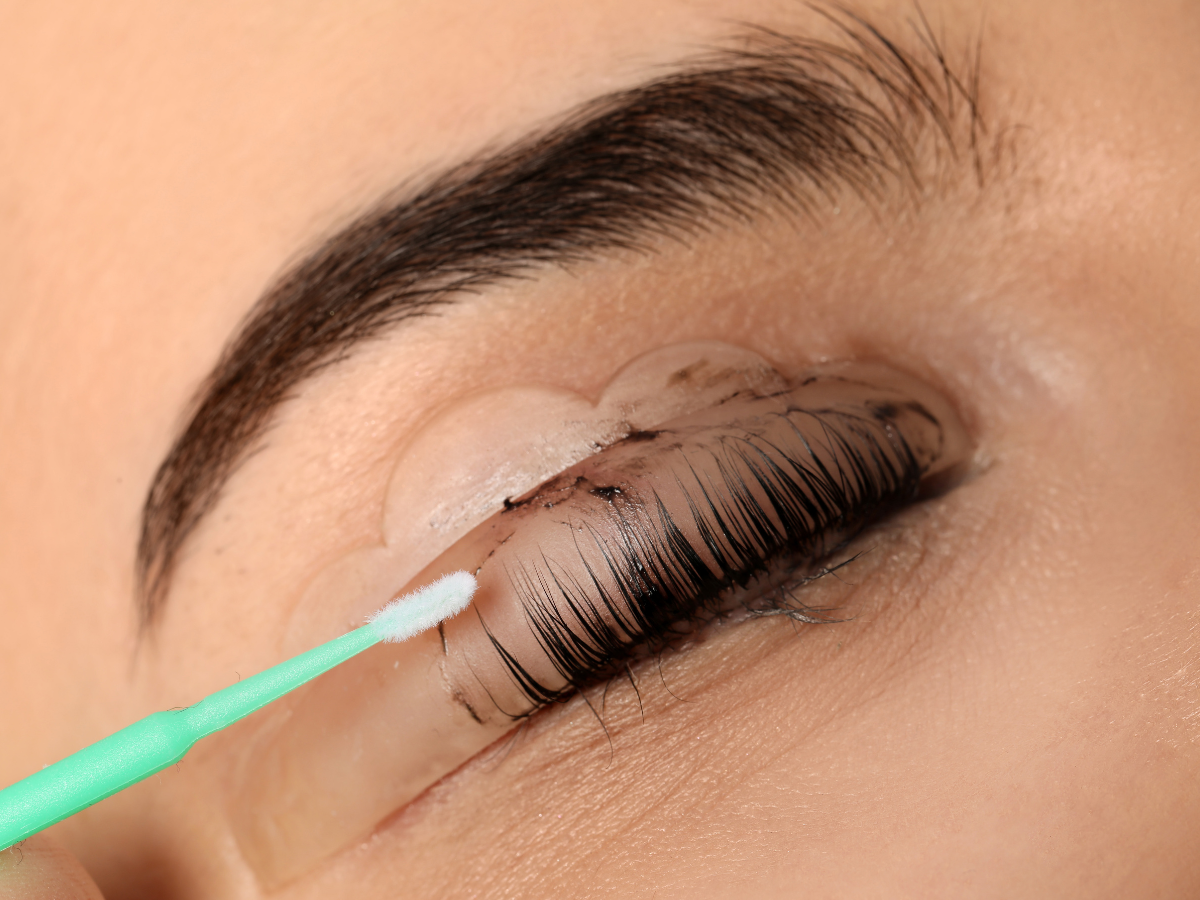 Eyelash lift procedure: Close-up of eye with lashes coated, being brushed with a small tool.