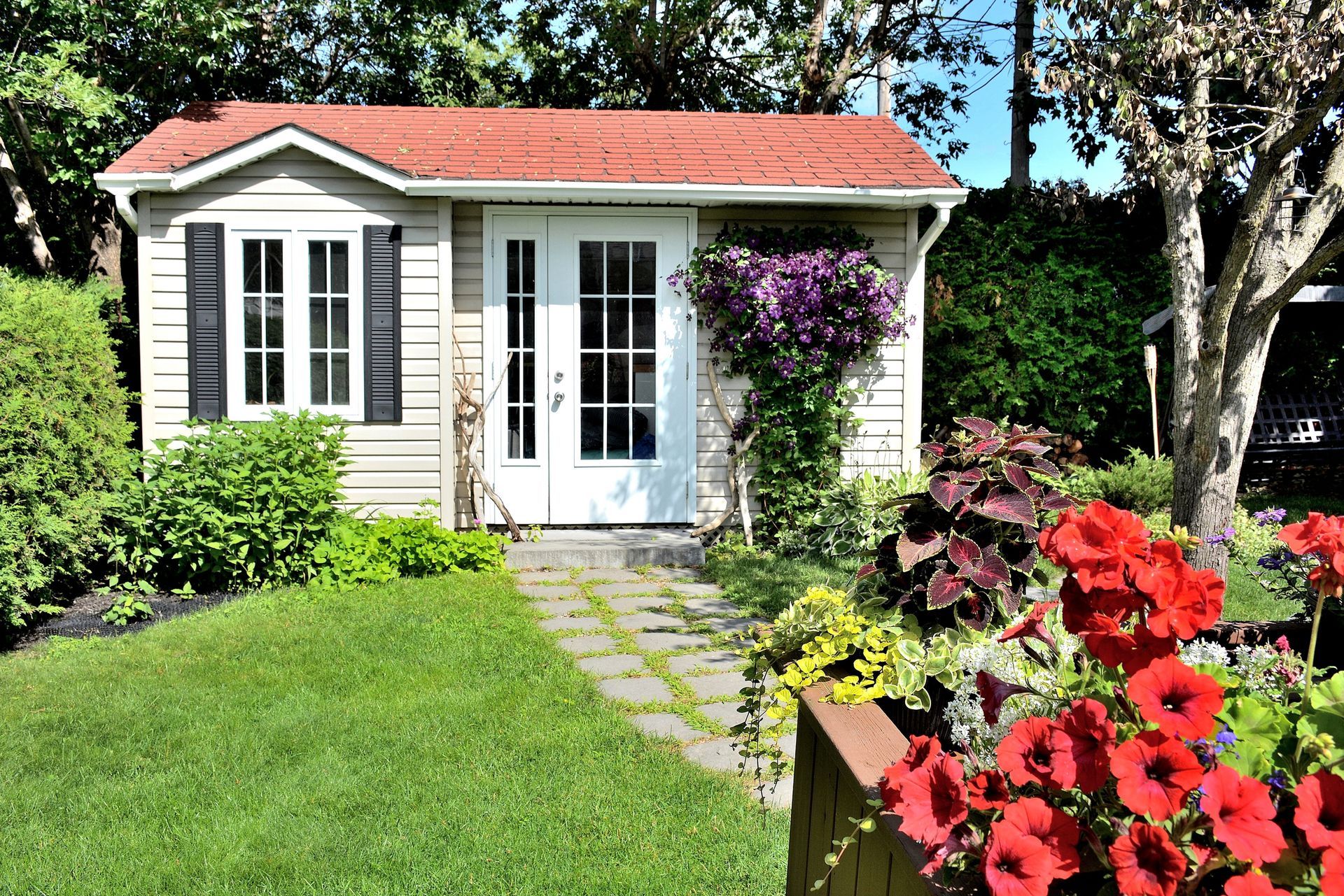 A small white house with a red roof is surrounded by flowers and trees.