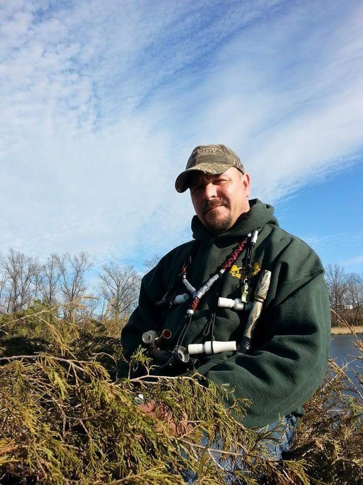 Man in camo hat and green jacket near brush and water under blue sky.