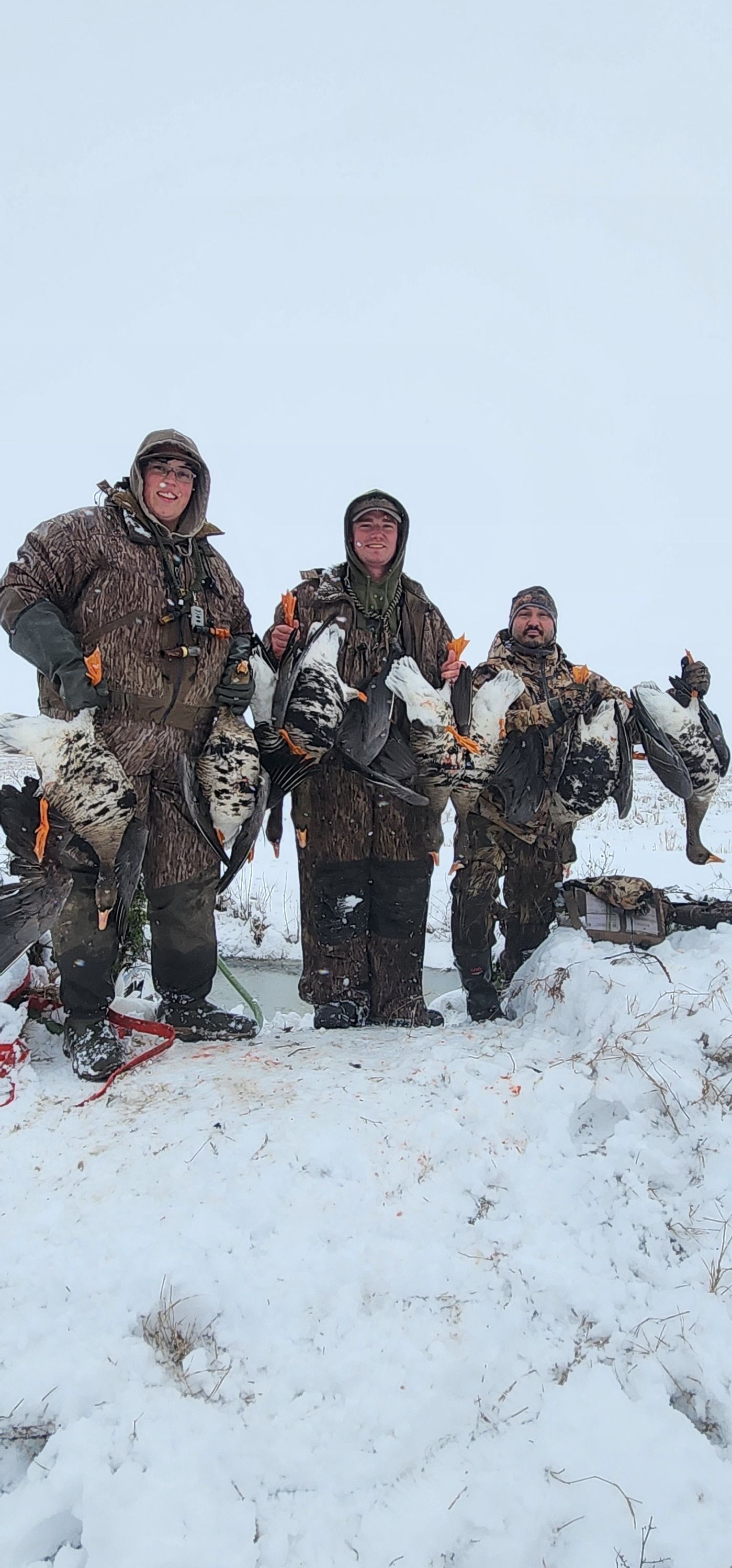 Three hunters in camouflage holding harvested ducks in a snowy field.