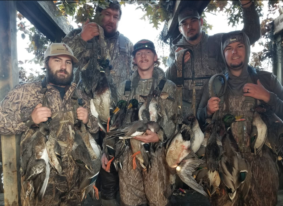 Group of hunters in camouflage, holding ducks they caught, smiling. Outdoor setting.