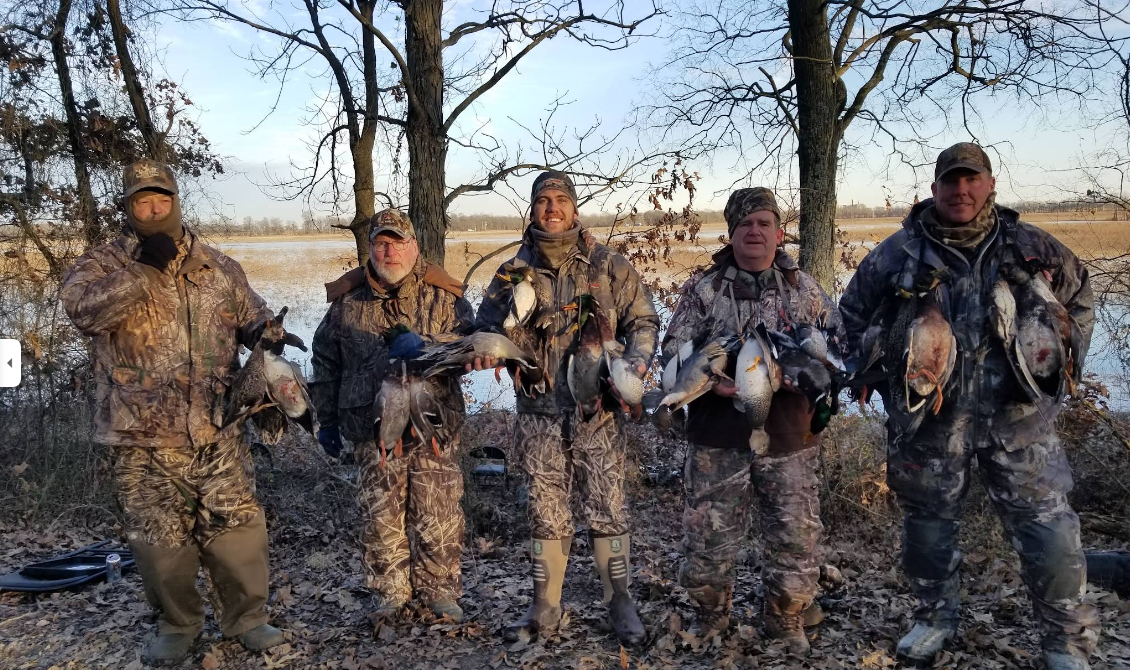 Two people stand beside a truck bed filled with hunted waterfowl on a muddy landscape under a bright, clear sky.