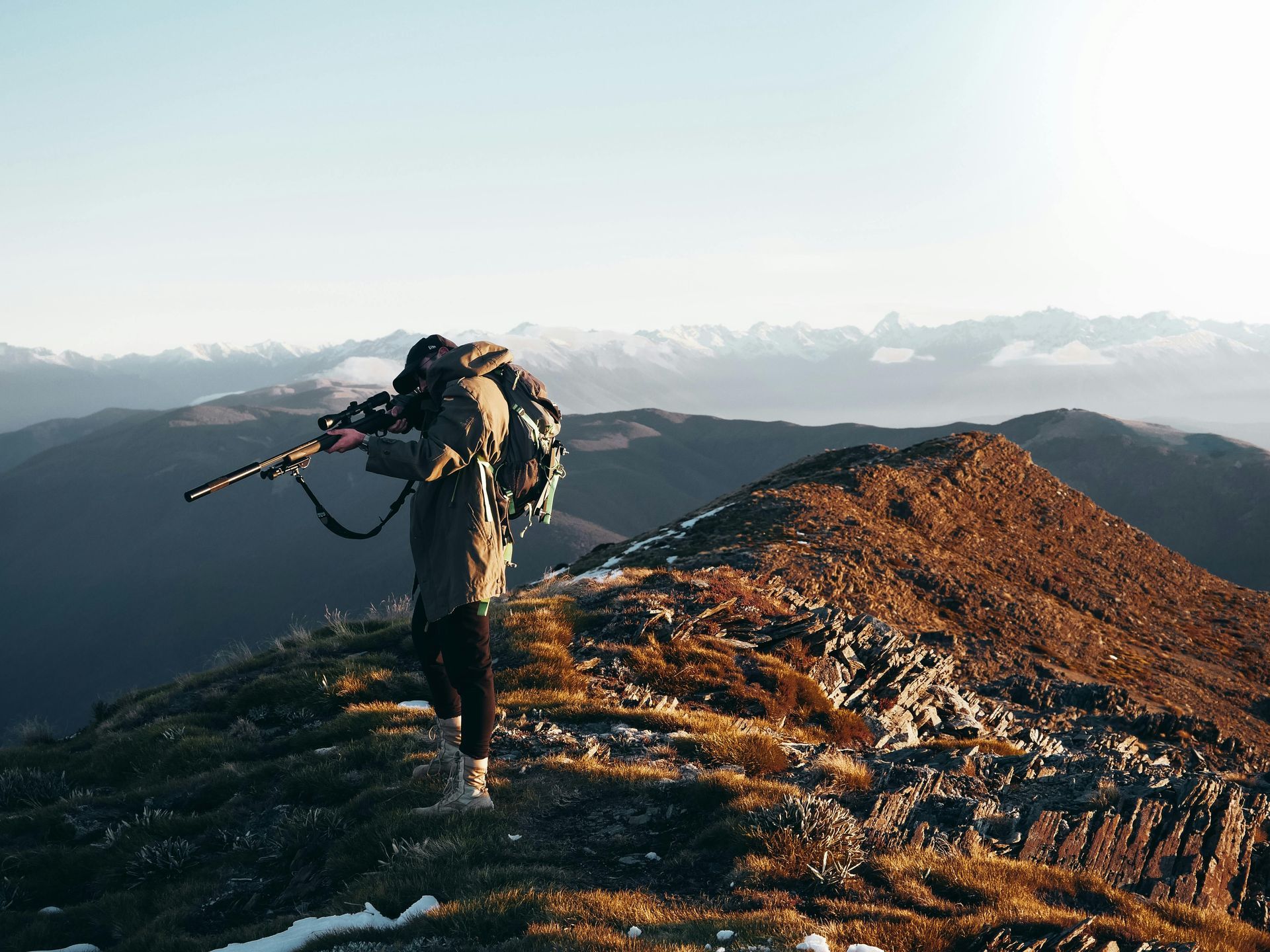 A person with a backpack stands on a mountain ridge at sunset, aiming a rifle toward the horizon.