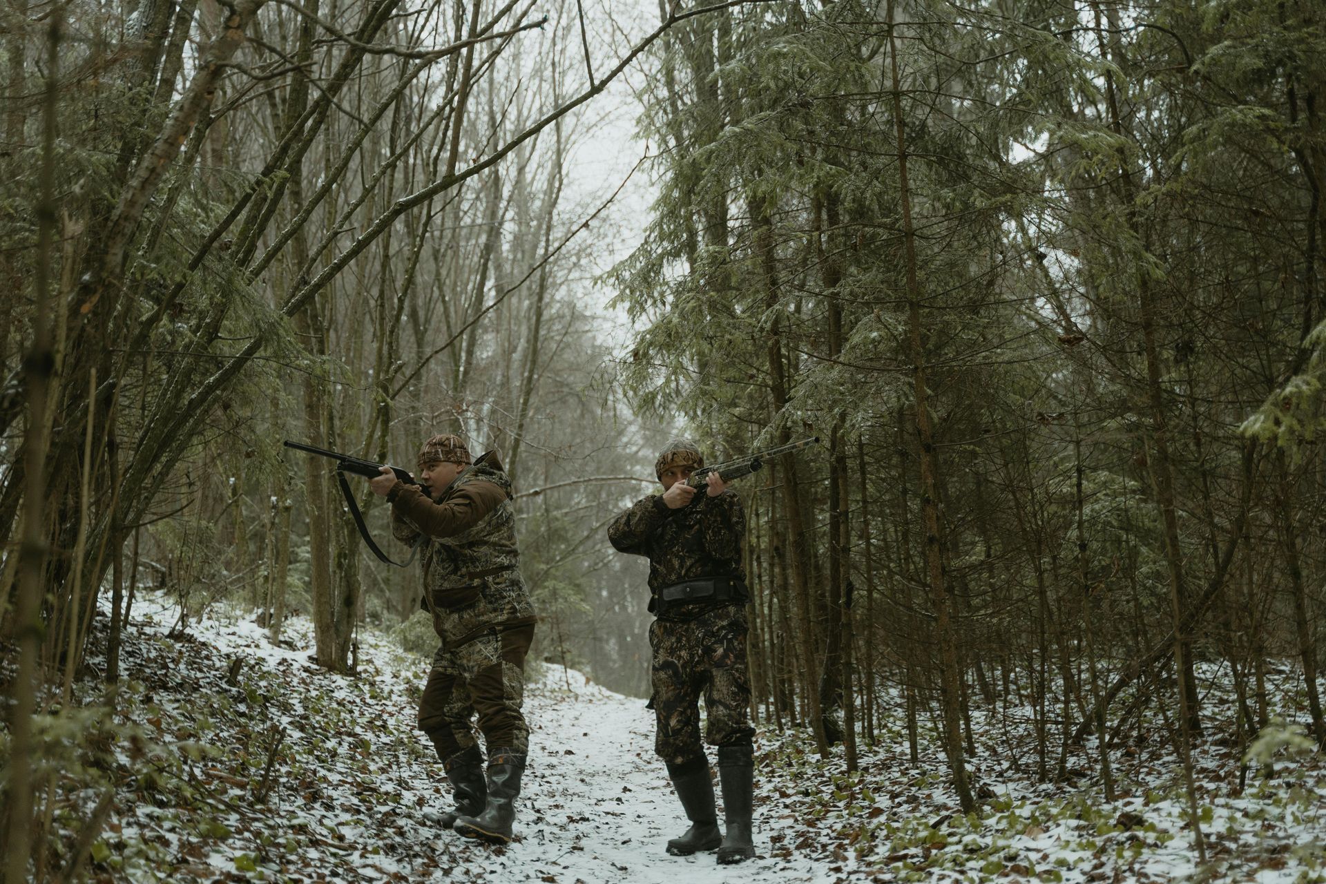 Two people in camouflage gear aim rifles while walking down a snow-covered path in a forest.