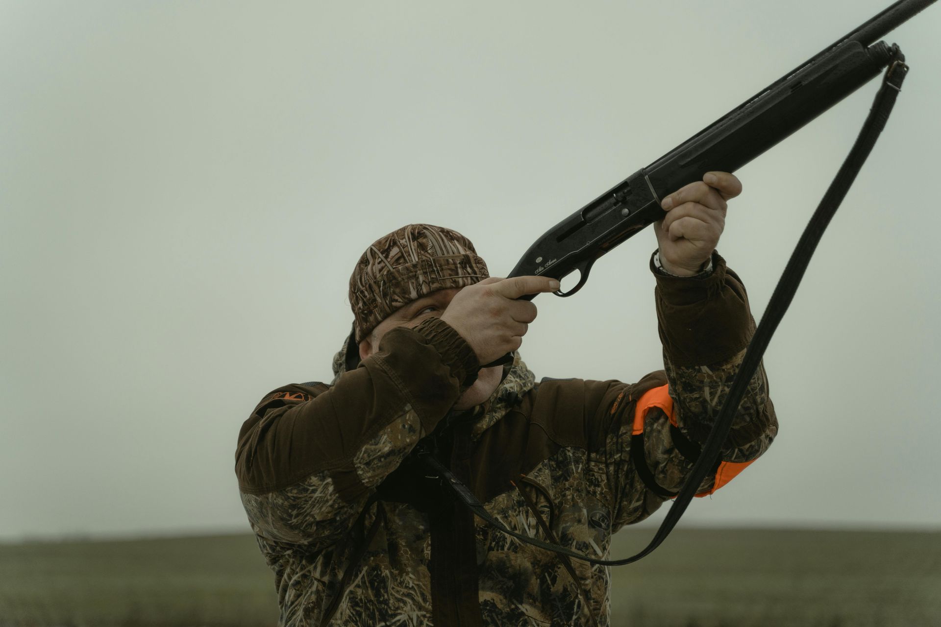 Hunter in camouflage aiming a shotgun outdoors under an overcast sky. Orange arm band visible.