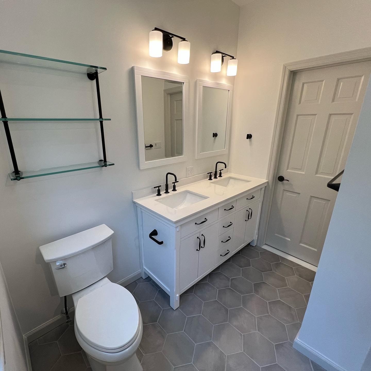 White bathroom with double vanity, mirrors, toilet, glass shelf, and door. Gray hexagonal floor tiles.