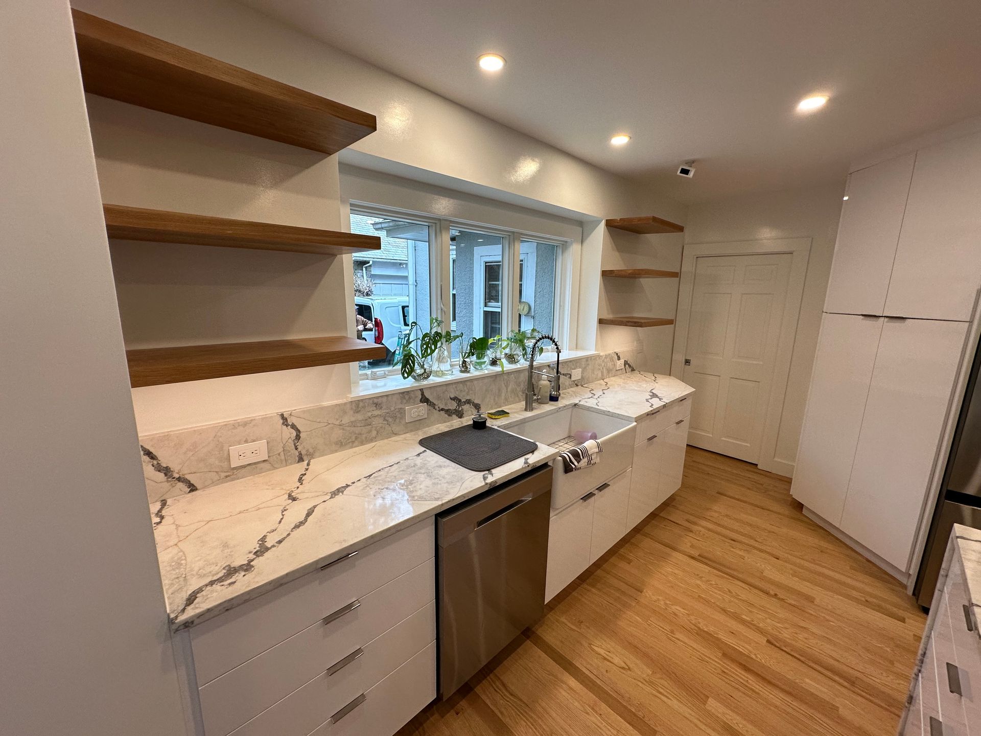 Modern kitchen with white cabinets, marble countertops, stainless steel appliances, and wooden shelves.