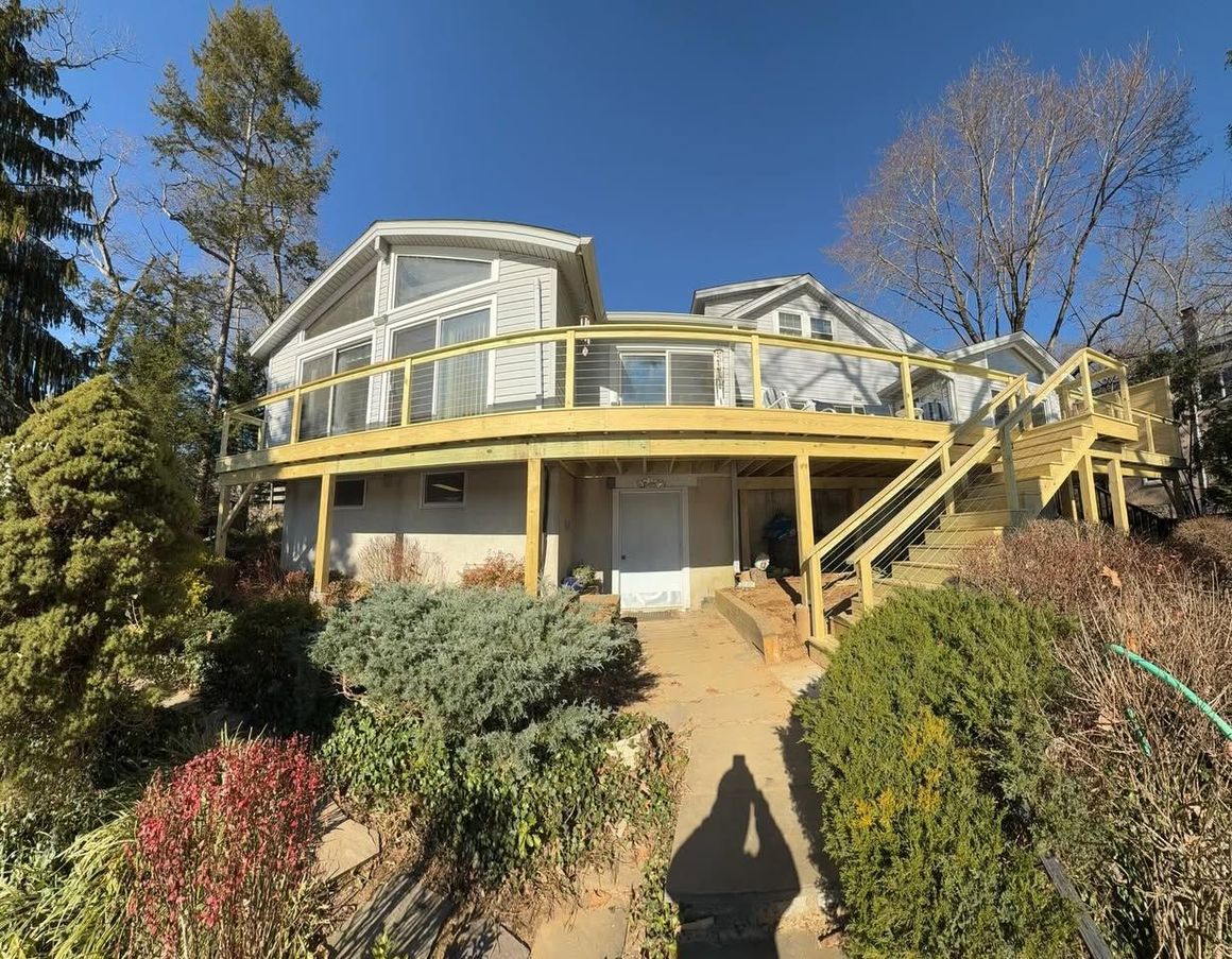 House with a large wooden deck. Exterior of house is light colored, surrounded by greenery and a clear blue sky.