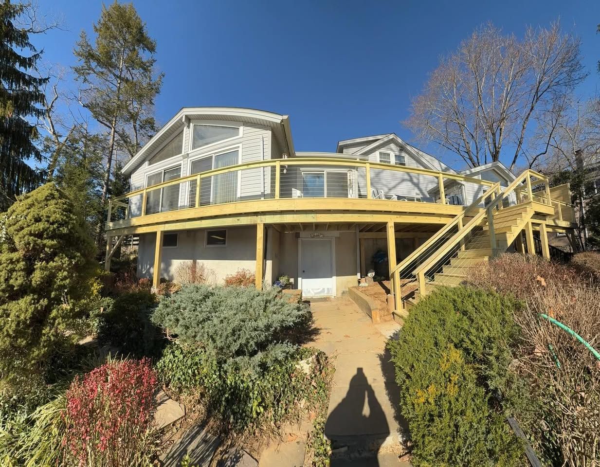 House with a large wooden deck. Exterior of house is light colored, surrounded by greenery and a clear blue sky.