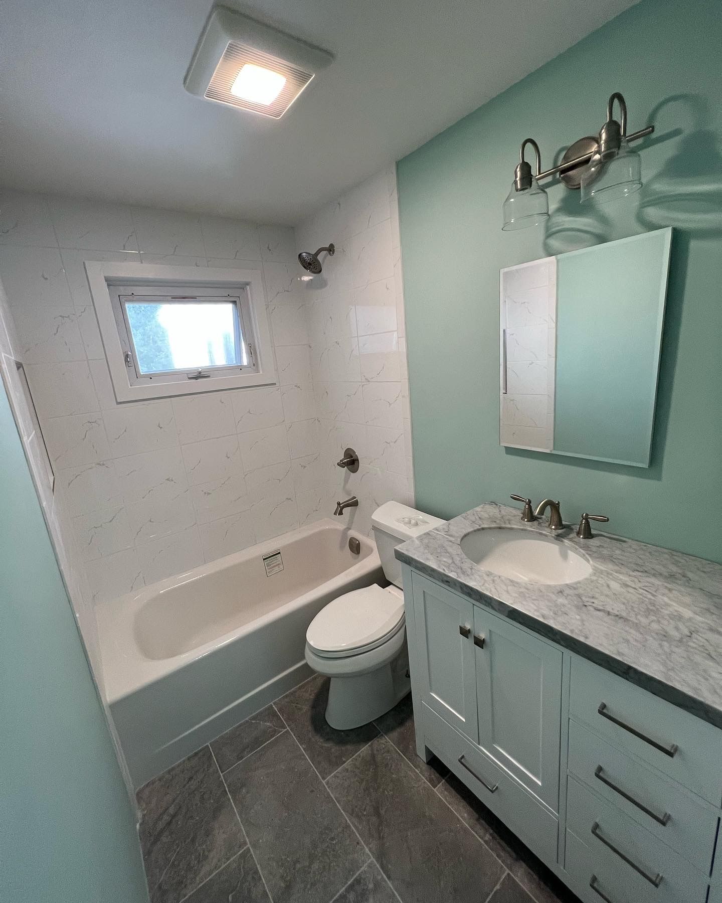 Small bathroom with teal vanity, marble counter, tub, toilet, and gray tile floor.