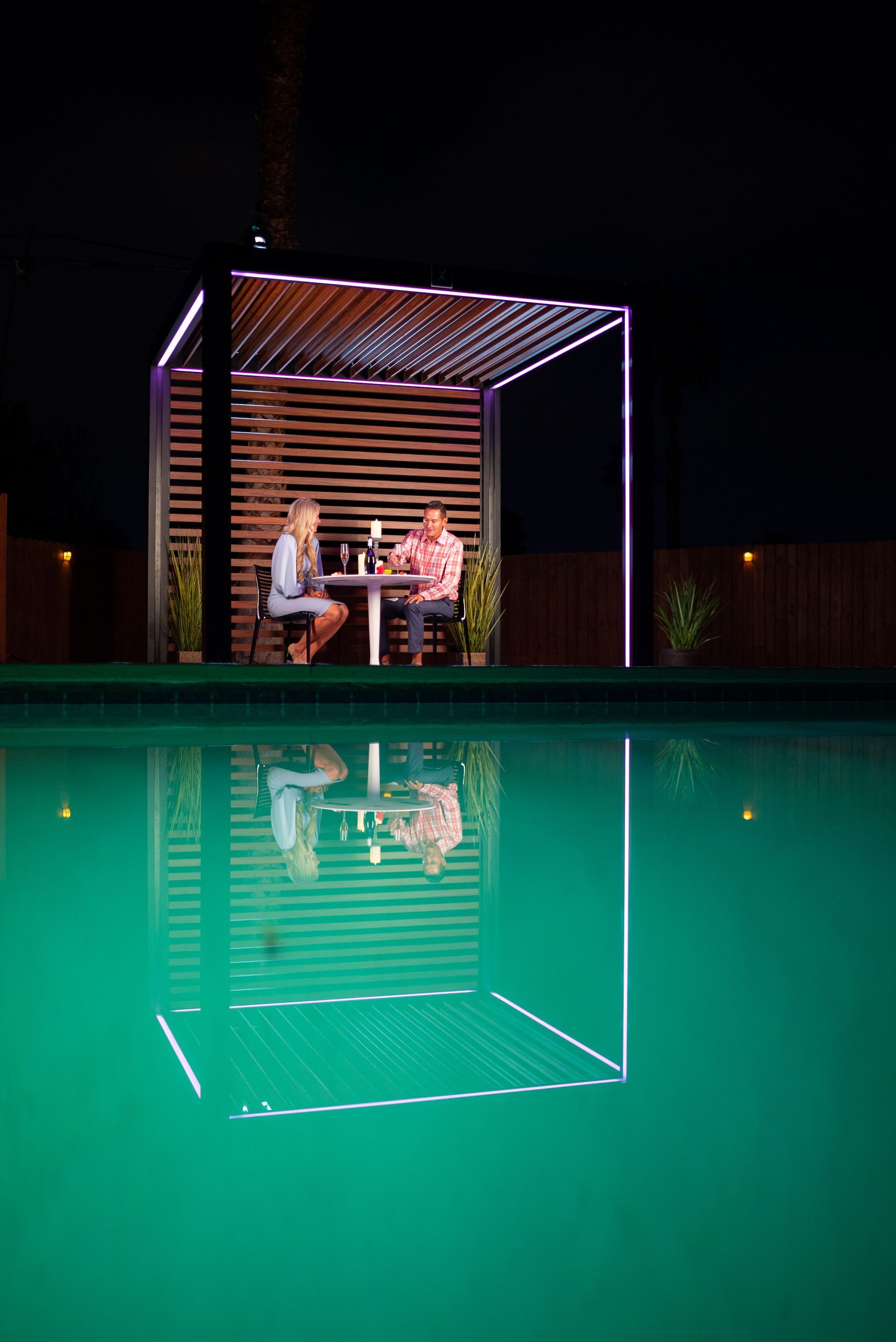 A couple is sitting at a table under a pergola next to a swimming pool at night.