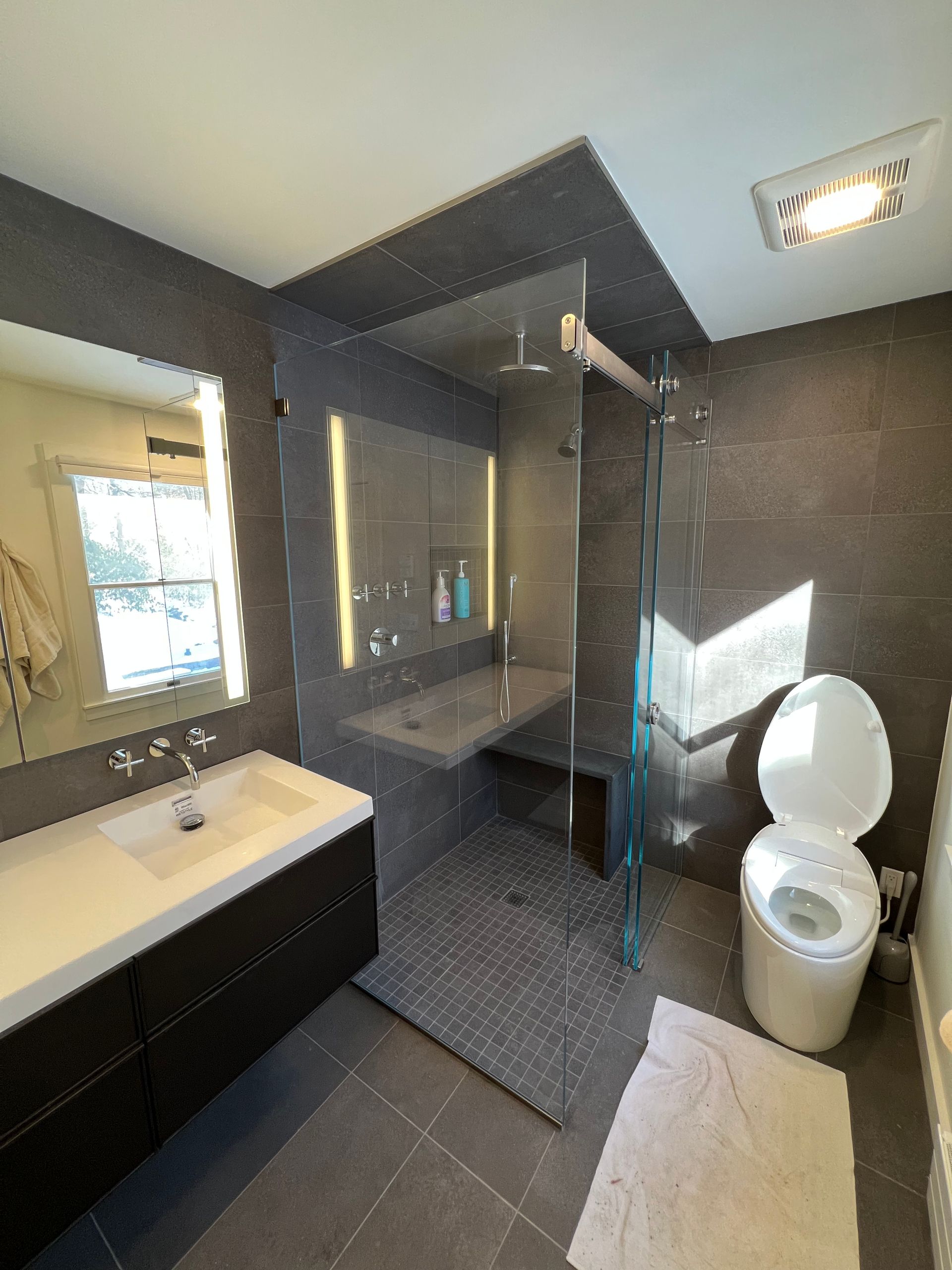 Modern bathroom with dark tile, glass shower, sink vanity, toilet, and window light