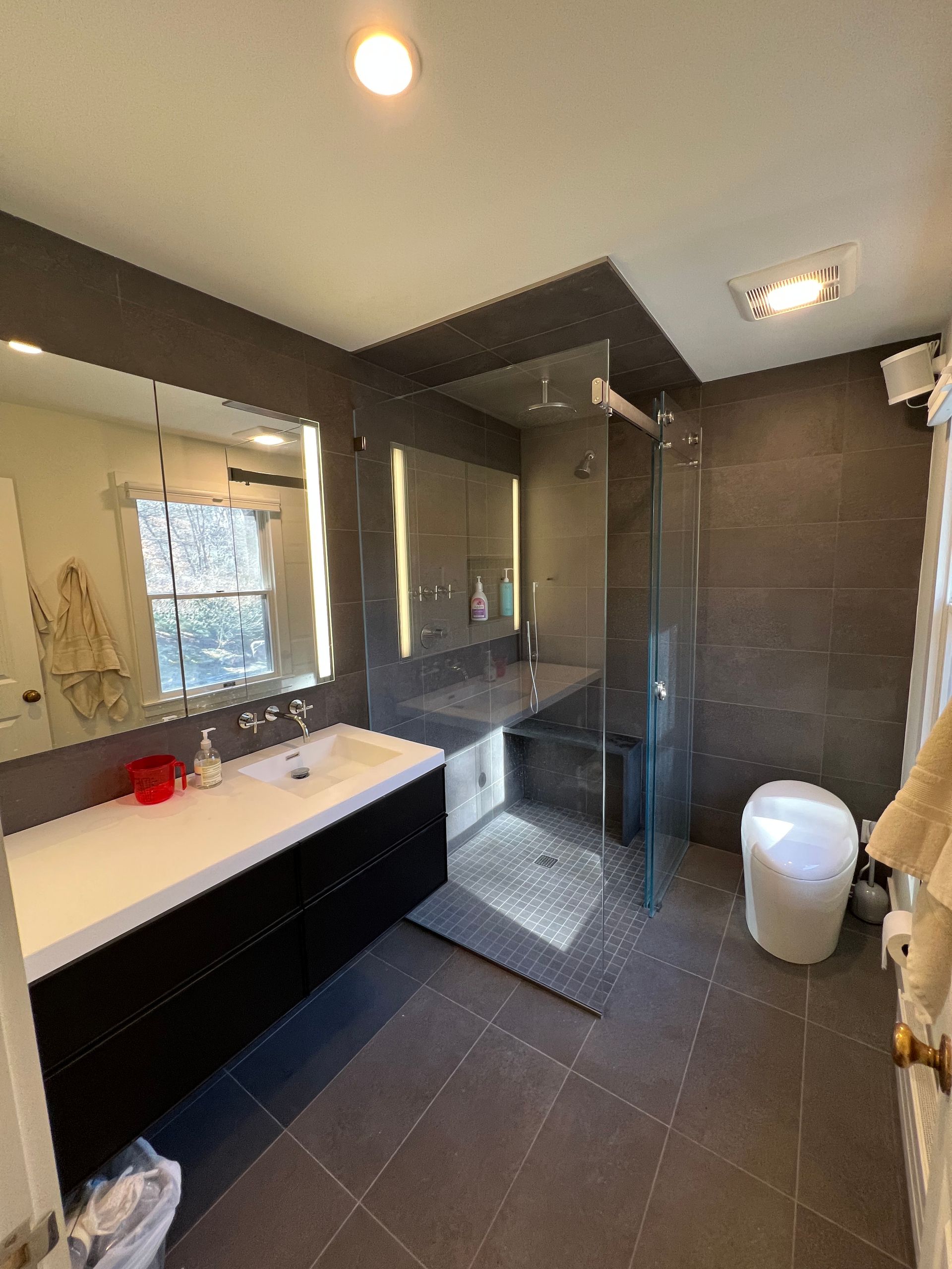 Modern bathroom with double sink, glass shower, dark tile, and skylight lighting