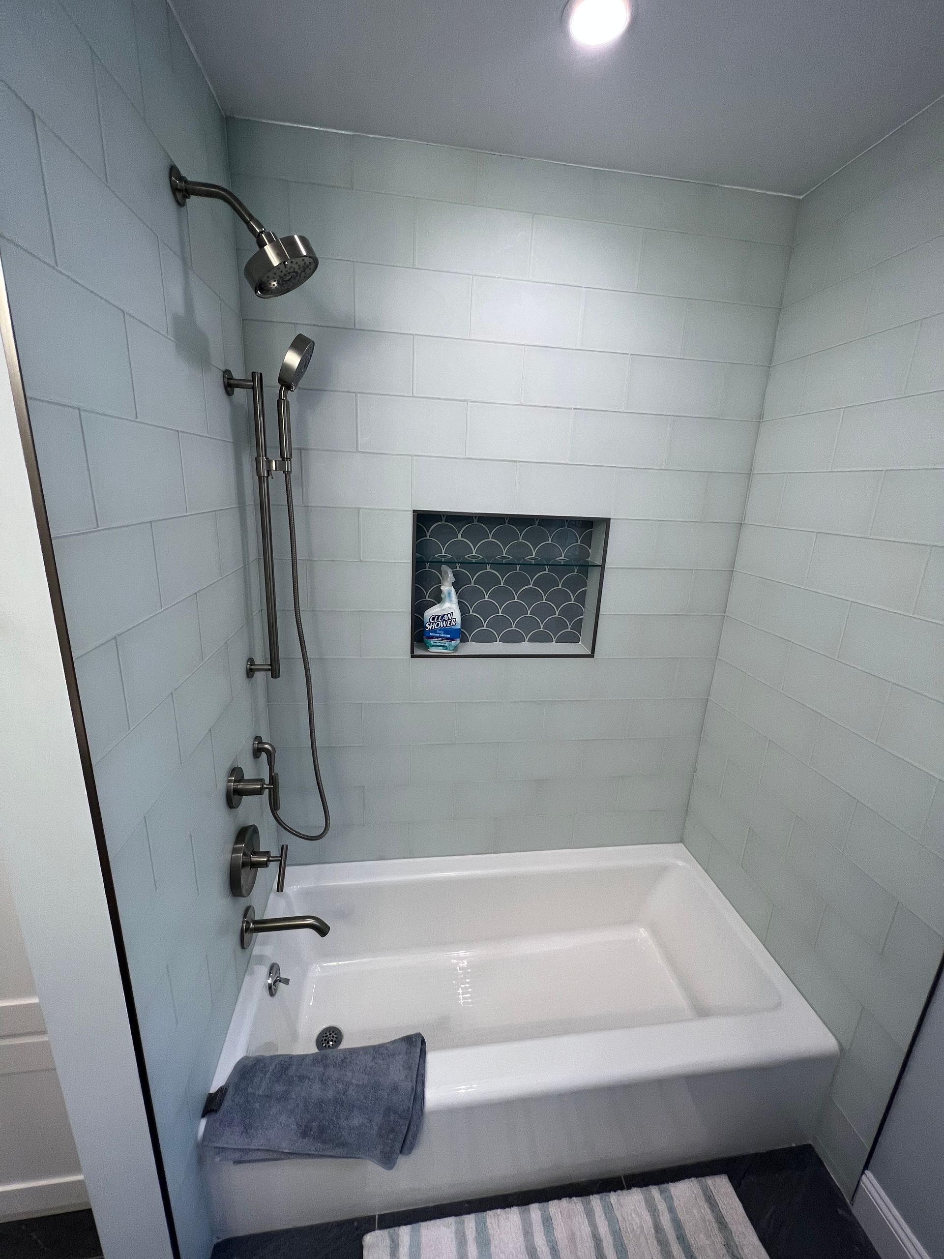 Small white tiled bathroom with a bathtub, showerhead, and towel on the rim.