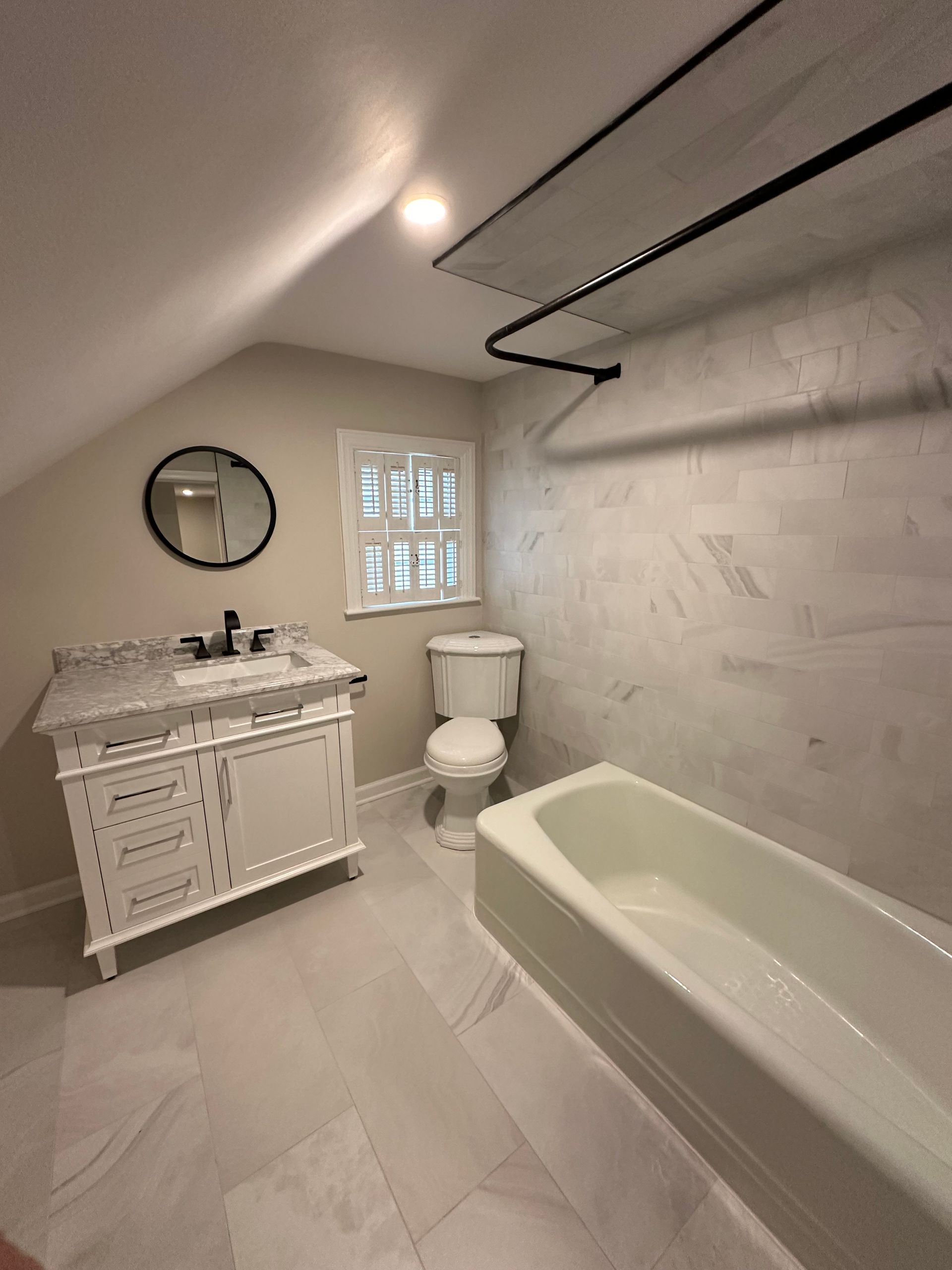 White bathroom with marble tub, toilet, ornate vanity, round mirror, and slanted ceiling
