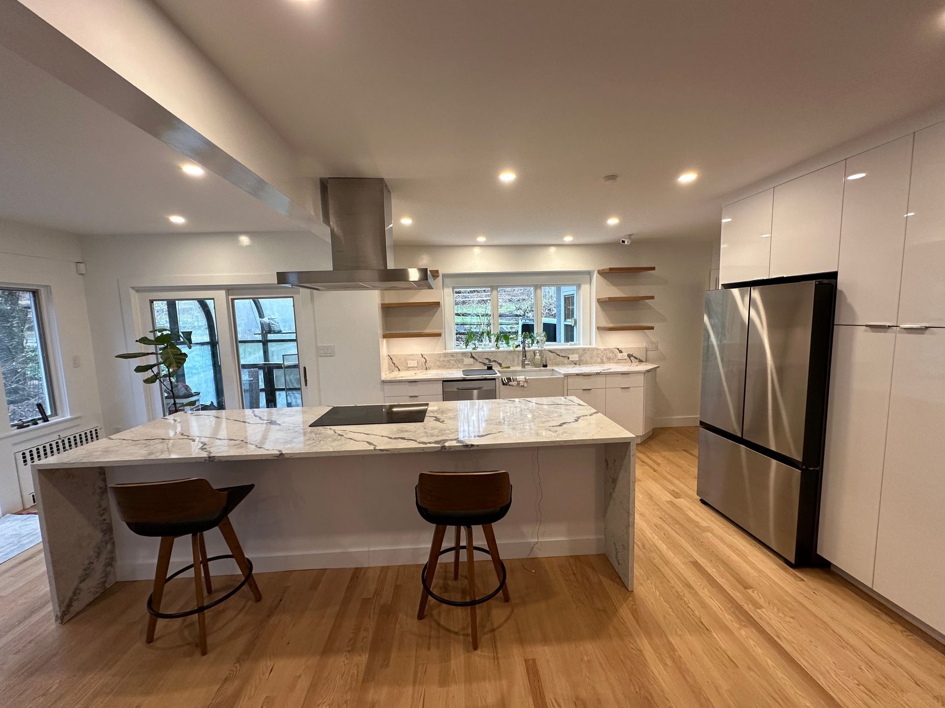 Modern white kitchen with marble island, two stools, stainless fridge, and bright windows