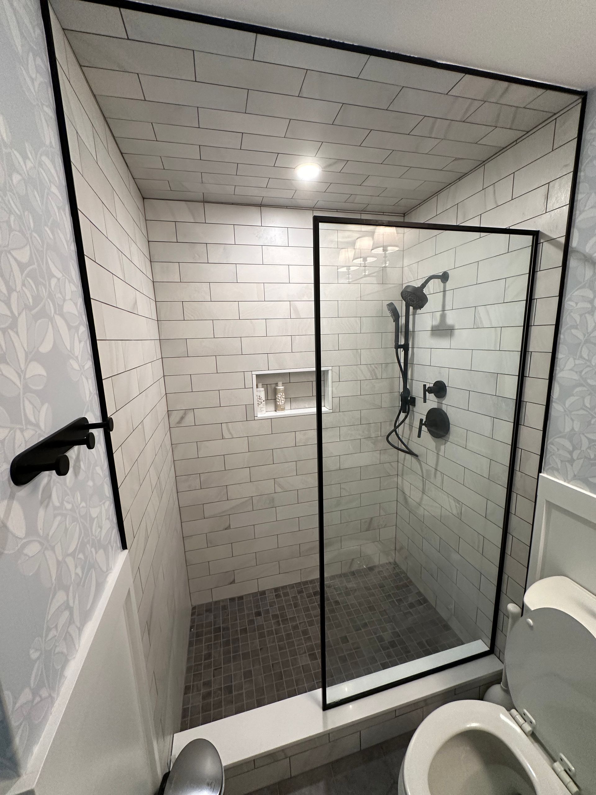 Modern tiled shower with glass door, built-in shelf, and rainfall showerhead in a bathroom.
