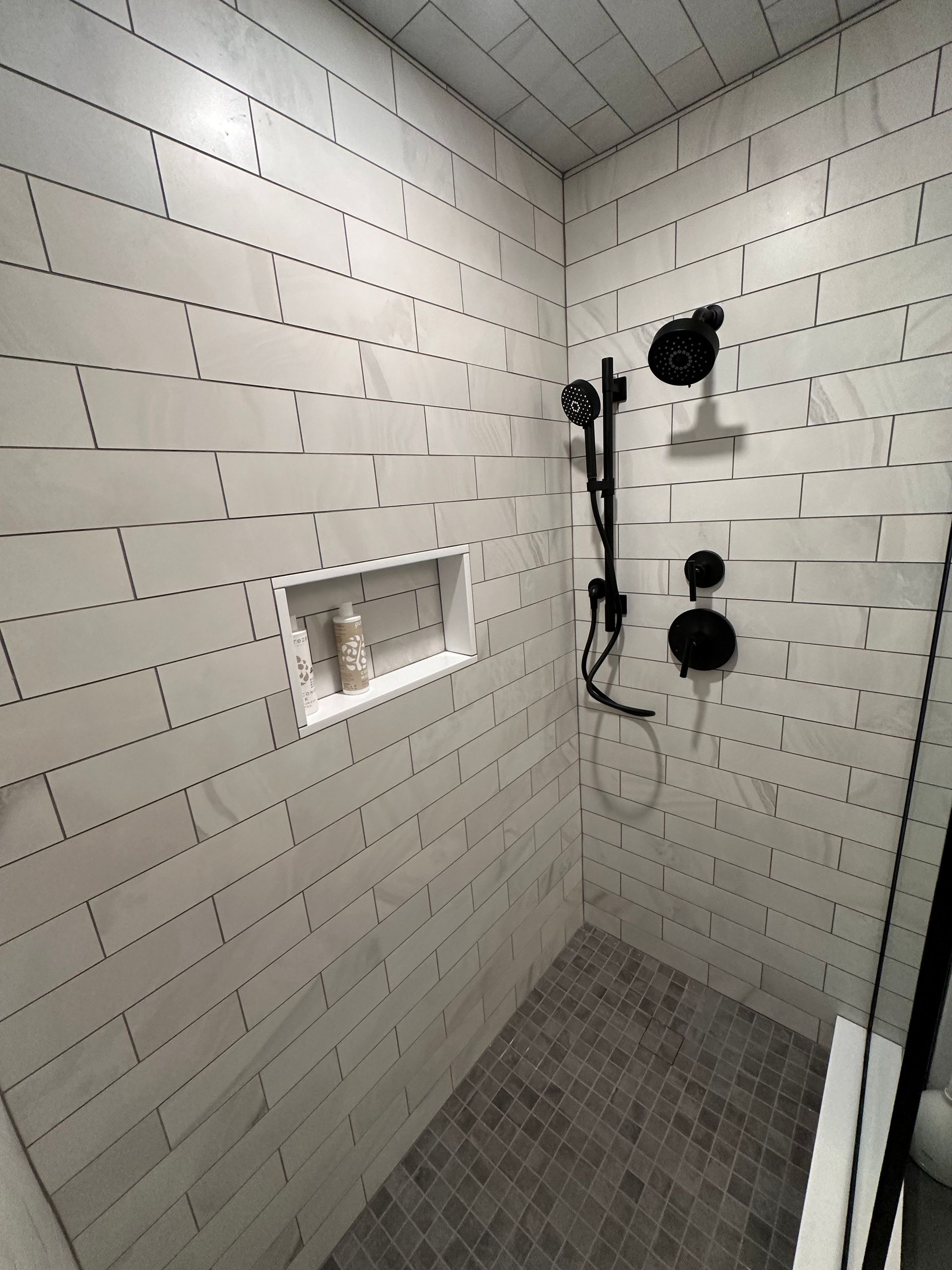 Empty tiled shower with two black shower fixtures and a recessed shelf.