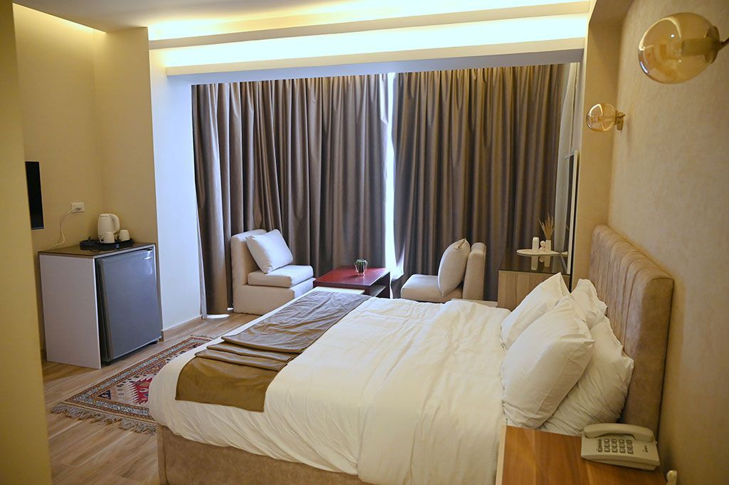 Hotel room with bed, chairs, curtains, and small refrigerator.