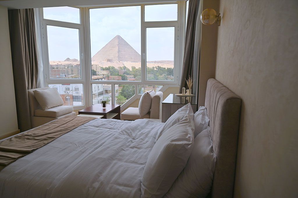 Bedroom with a large window overlooking the Great Pyramid of Giza; bed with white linens, two chairs, and a small table.