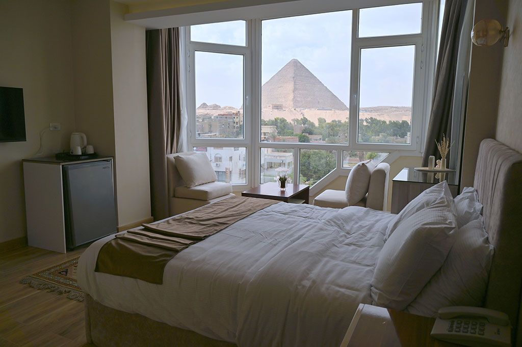 Bedroom with large window overlooking a pyramid in Egypt. Bed, chairs, and a small refrigerator are visible.