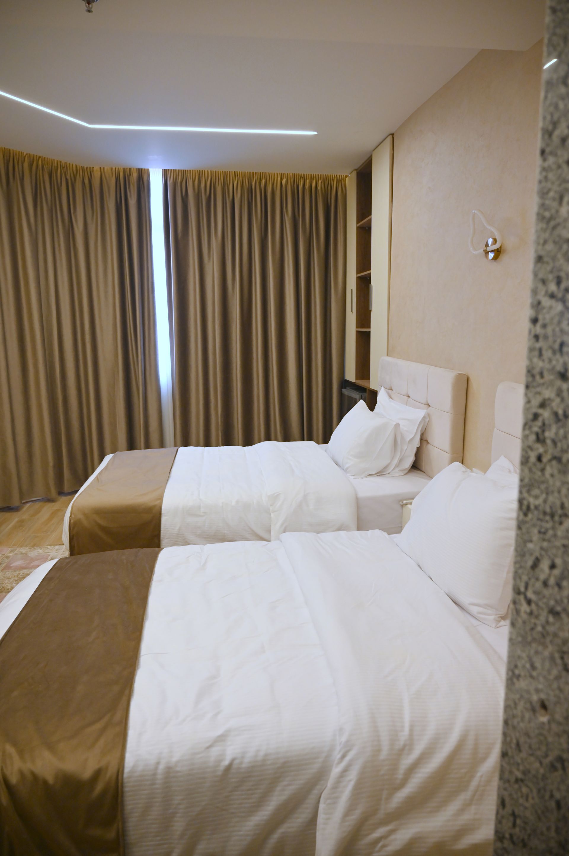 Two twin beds in a hotel room with tan and white bedding, with curtains and a wall fixture.