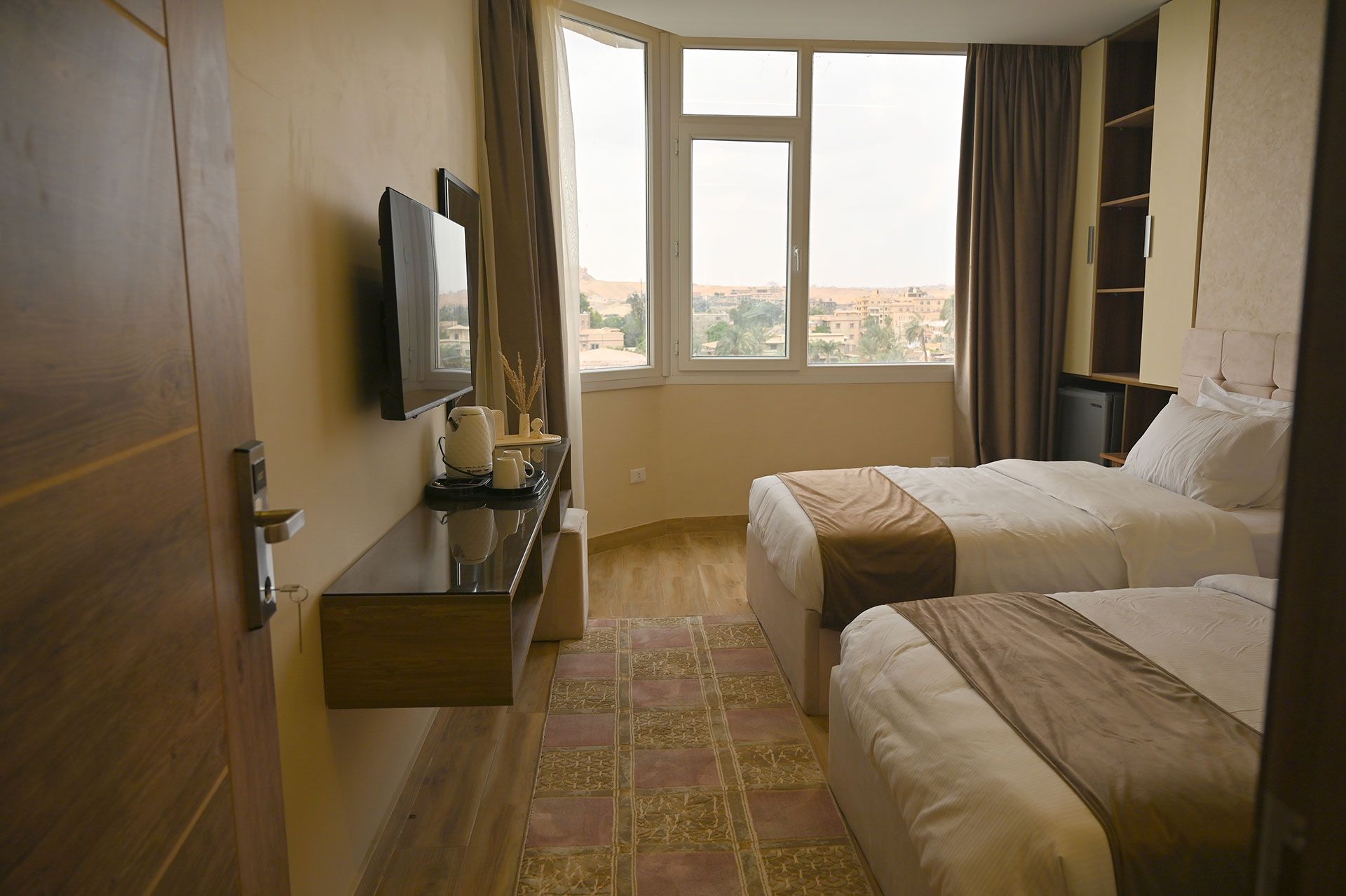 Hotel room with two beds, TV, window, and a door on a light brown wall.